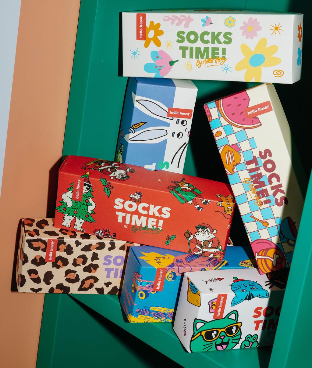 Cofanetti regalo "Socks Time !" impilati su una mensola verde, packaging colorati e illustrati: motivi Natale, fiori, leopard, mascotte, auto, gatti e cibo – Hello Hossy