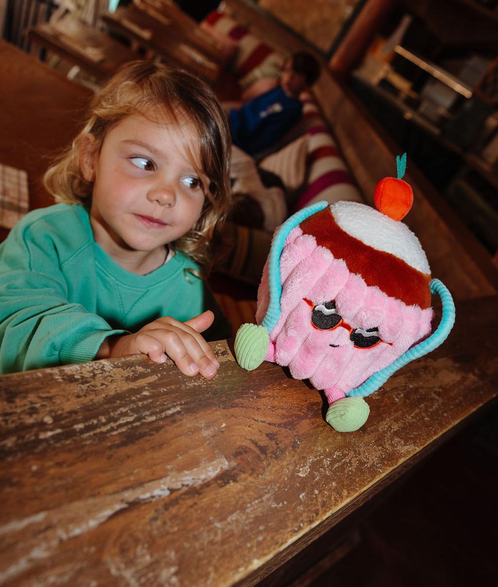 Peluche a forma di torta con occhiali da sole Cherry Cake, posata su una rampa con un bambino che la guarda - giocattolo originale - Hello Hossy