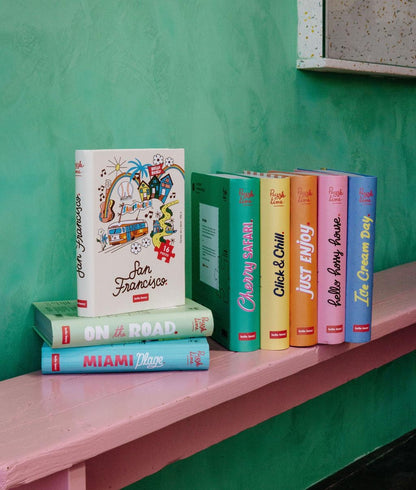 Collezione di puzzle Hello Hossy posati su una panca rosa – design effetto libro – sfondo parete colorato