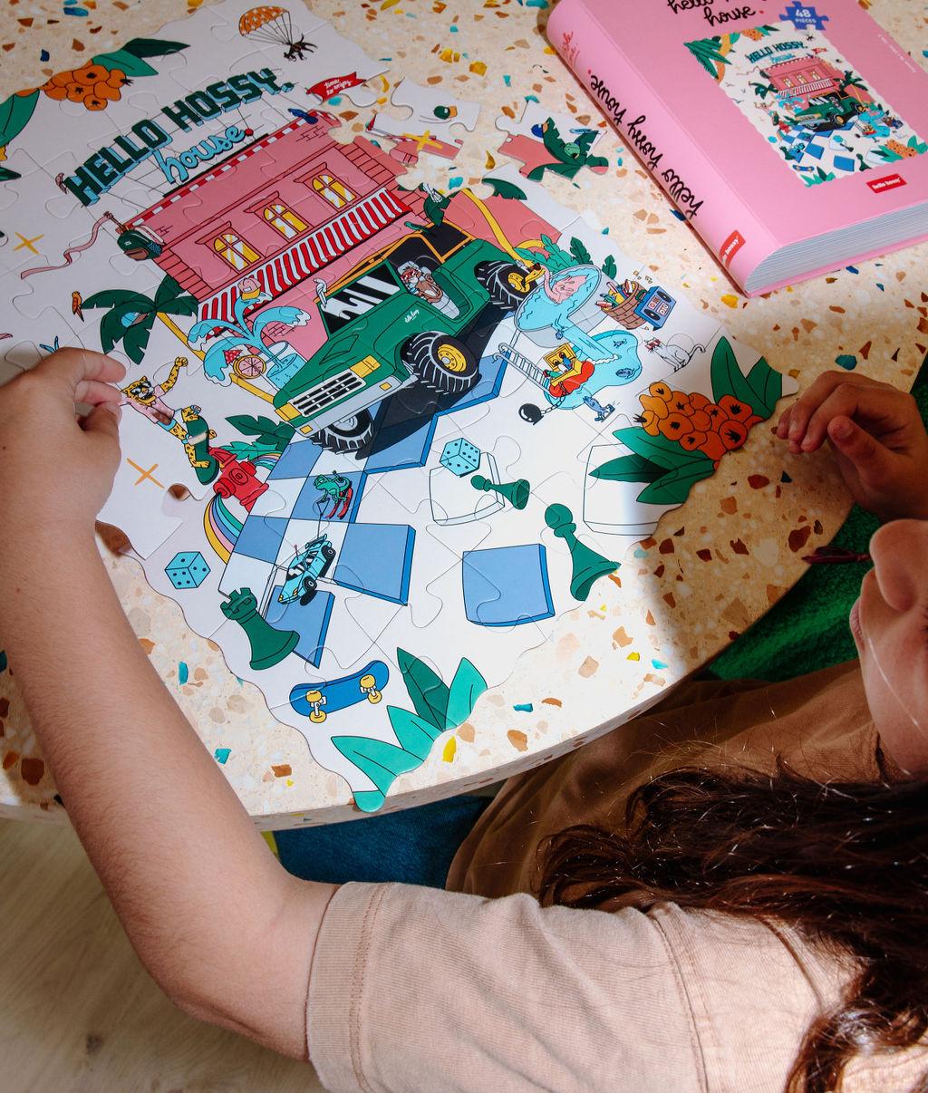 Bambina che compone il Puzzle colorato da 48 pezzi "Hello Hossy House" su un tavolo – vista dall'alto – Hello Hossy