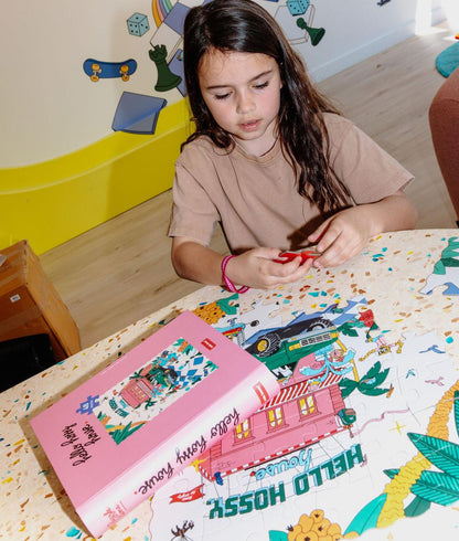 Bambina che compone il Puzzle illustrato "Hello Hossy House" su un tavolo, con la scatola accanto – foto lifestyle – Hello Hossy