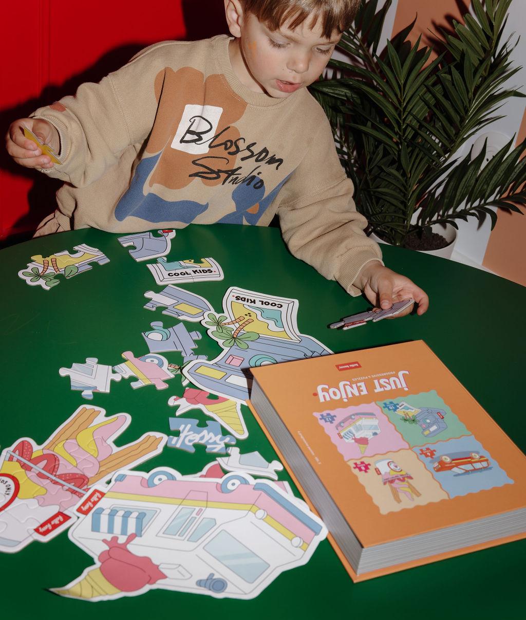 Bambino che assembla i puzzle colorati "Just Enjoy" su un tavolo, con i modelli davanti a sé – atmosfera giocosa – Hello Hossy