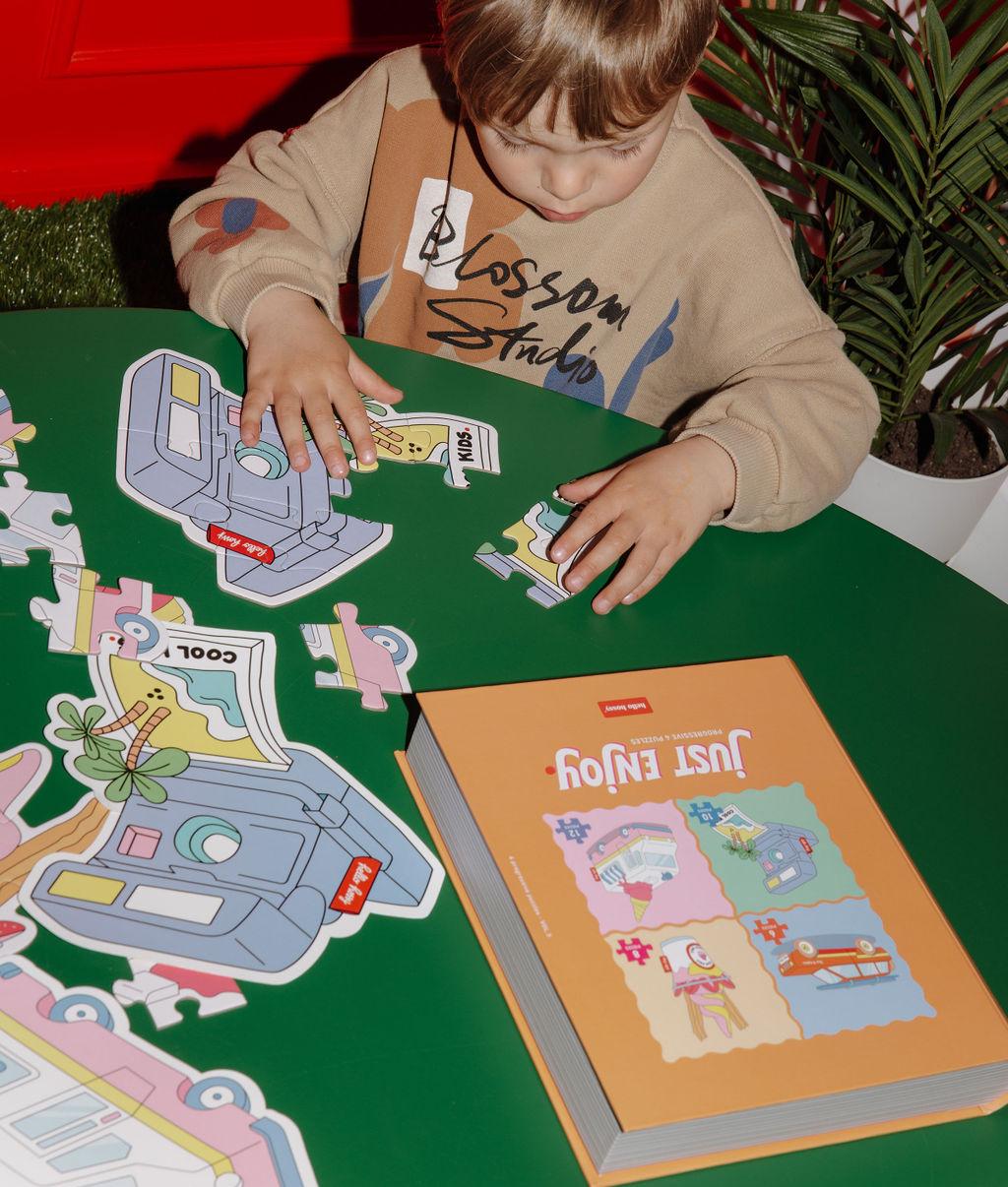 Bambino con uno dei quattro puzzle "Just Enjoy" parzialmente assemblato, la scatola e l'illustrazione modello, su un tavolo davanti a lui – foto lifestyle – Hello Hossy