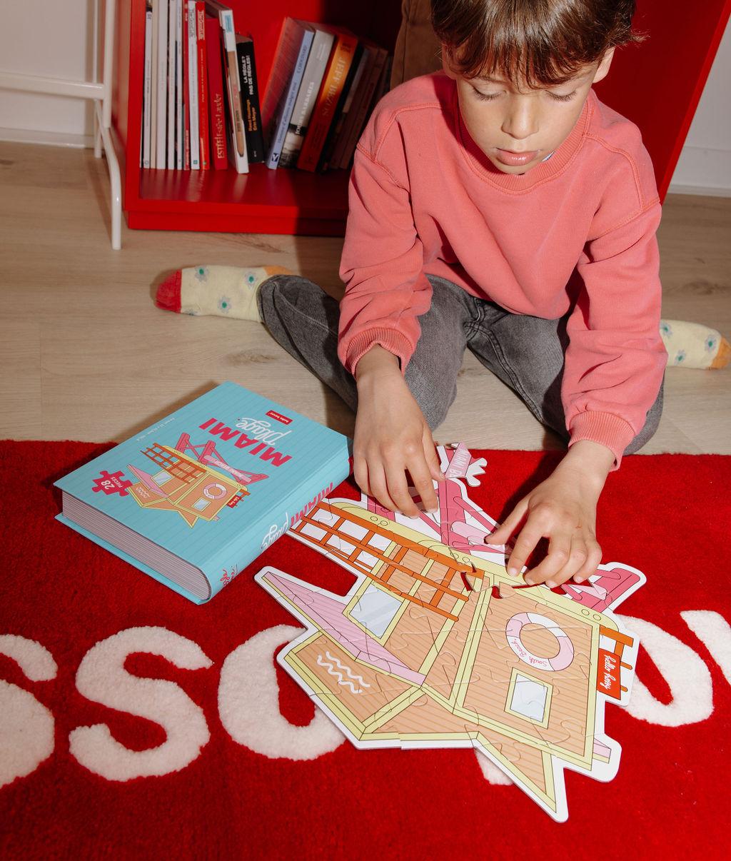 Bambino con il Puzzle "Miami Plage" parzialmente assemblato e la scatola, su un tappeto, davanti a lui – foto lifestyle – Hello Hossy