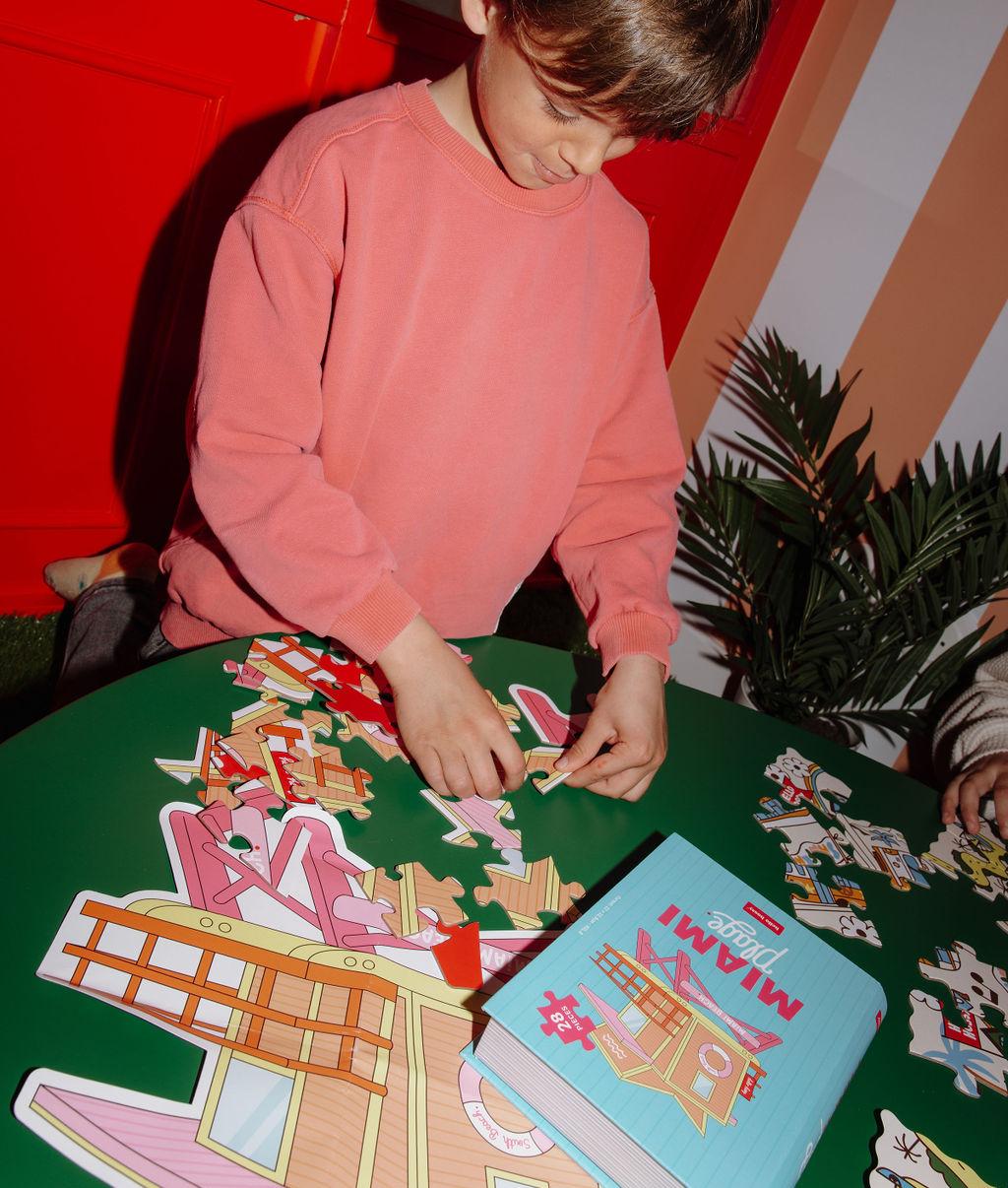 Bambino che gioca con il Puzzle 28 pezzi "Miami Plage" su un tavolo con la scatola accanto – atmosfera giocosa – Hello Hossy