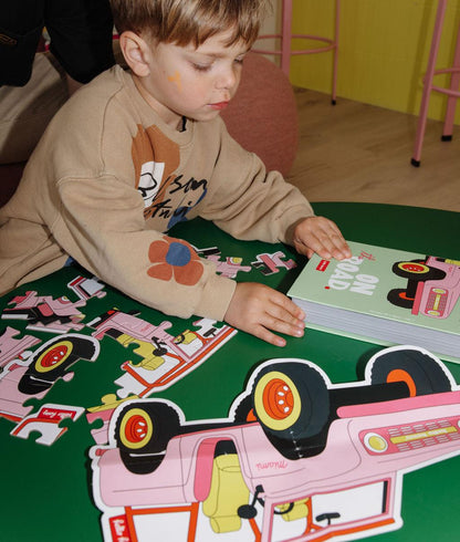 Ragazzo con il puzzle "On The Road" parzialmente assemblato, la scatola e l'illustrazione modello, su un tavolo davanti a lui – foto lifestyle – Hello Hossy